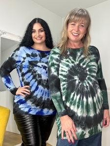 Two women smile and pose indoors, showcasing comfort and style. One wears a blue tie-dye Tally Jumper with black pants, the other in a green version with jeans. Their super-soft ensembles embrace the cozy knit trend as they stand against a light wall with a mirror behind them.