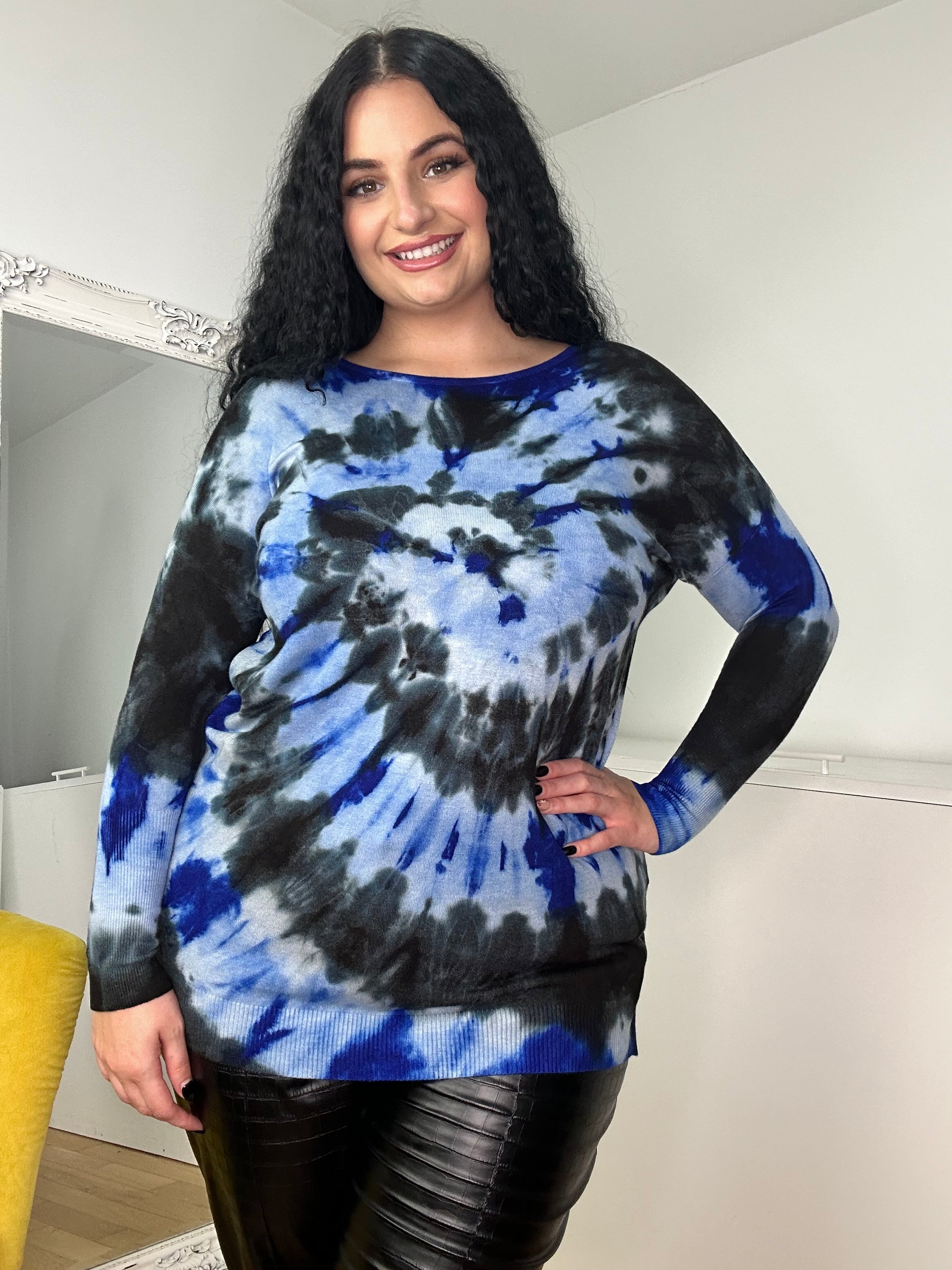 A person with long, curly hair beams while posing in a room, wrapped snugly in the ultra-soft Tally Jumper featuring a blue and black tie-dye design, paired with black leather pants. A mirror and part of a yellow chair add charm to the scene.