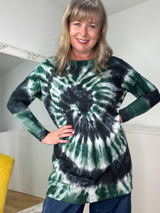 A woman smiles and poses indoors in the super soft, one-size-fit Tally Jumper tie-dye shirt in green, black, and white. She stands confidently with hands on hips, her shoulder-length hair framing her face. A yellow chair is partially visible to the left.