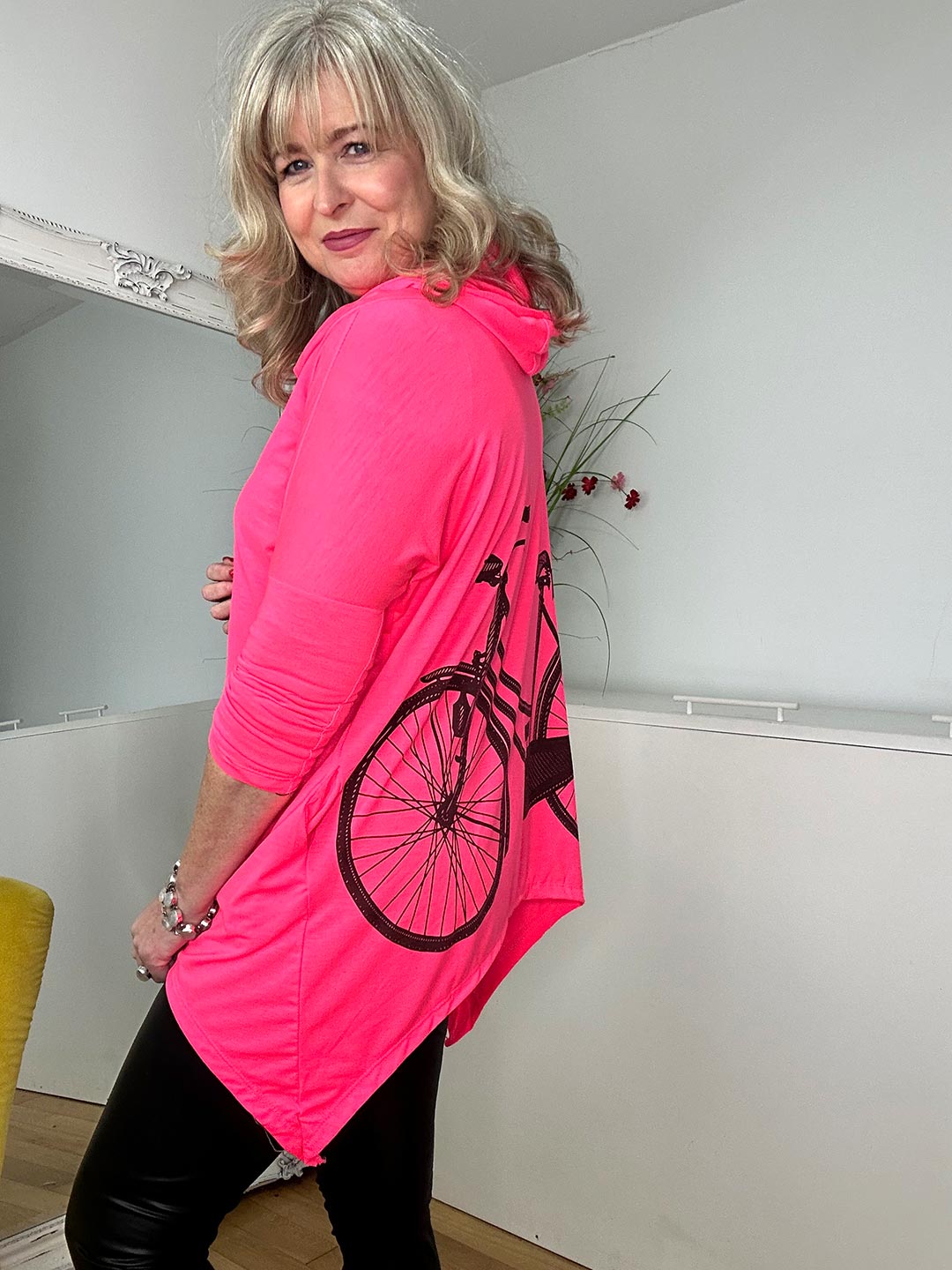 A woman in a bright pink top and black leggings stands in a cozy room, smiling confidently.