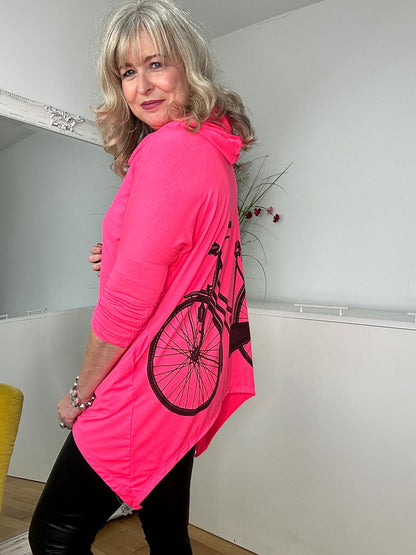 A woman in a bright pink top and black leggings stands in a cozy room, smiling confidently.