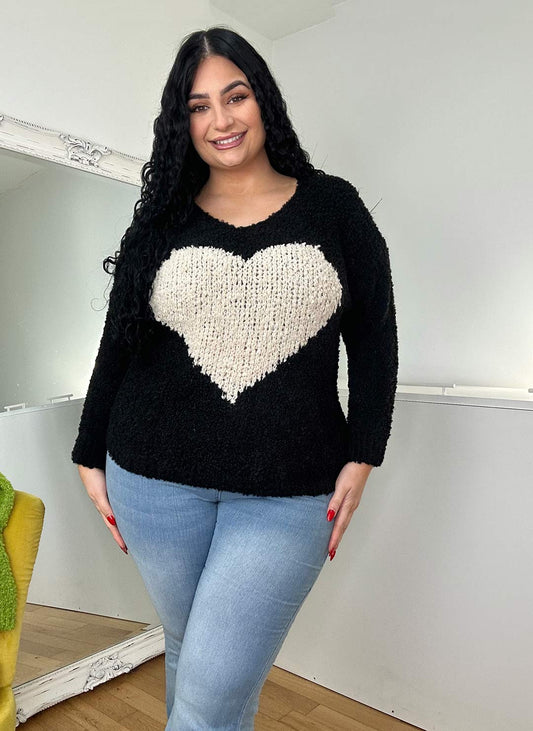 A woman with curly black hair wearing a black sweater with a large cream heart design and light blue jeans, standing confidently in a well-lit room.