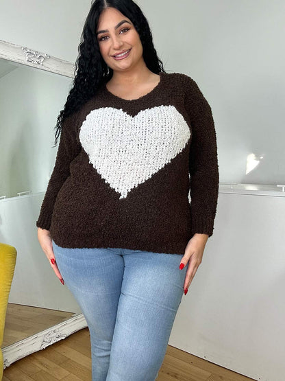 A woman with curly black hair wearing a dark brown sweater with a white heart design and light blue jeans, smiling warmly, standing in a bright room.