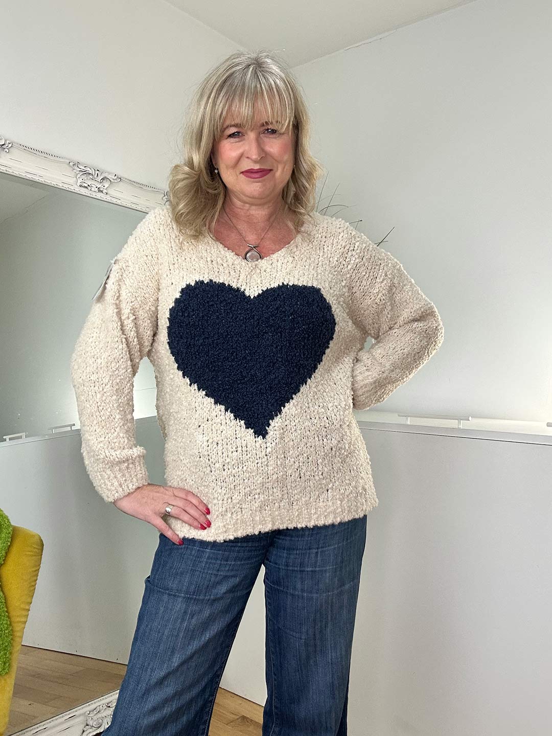 A woman with blonde hair wearing a cream-colored sweater with a navy-blue heart design and denim jeans, posing casually in a cozy indoor setting.