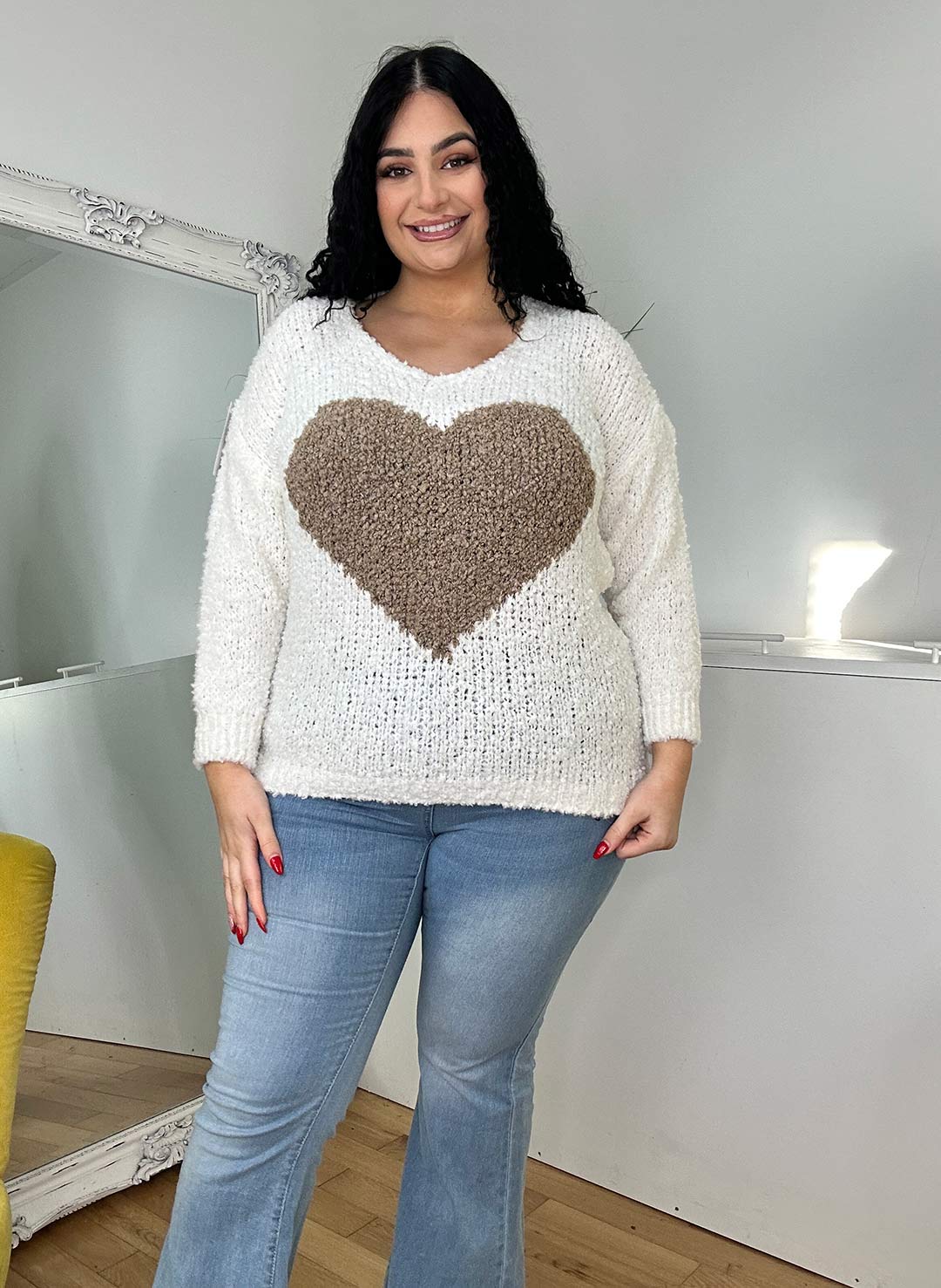 A woman with curly black hair wearing an ivory sweater with a tan heart design and light blue jeans, standing cheerfully in a minimalistic room.