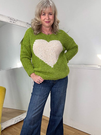 A woman with blonde hair wearing a vibrant green sweater with a cream heart design and denim jeans, smiling softly in a bright and cozy space.