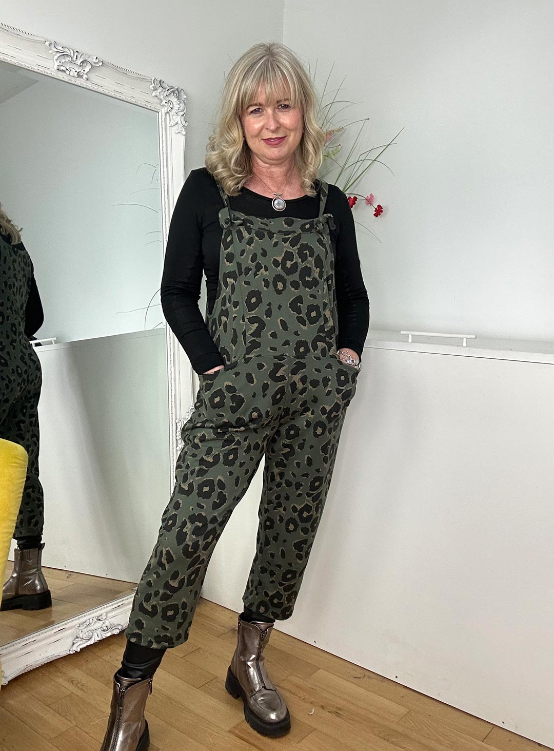  A woman in khaki leopard print dungarees stands confidently in front of a mirror, reflecting her stylish and casual look.