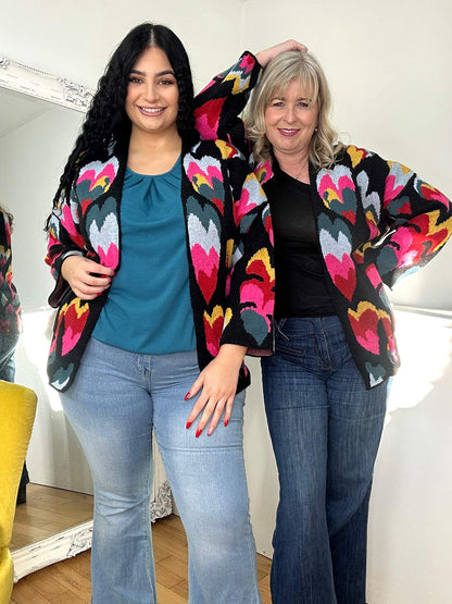 Two women, one with curly black hair and one with blonde hair, wearing matching vibrant cardigans with colorful heart patterns