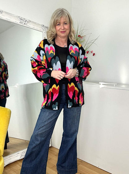 A woman is standing indoors, wearing a colorful cardigan with a pattern of heart-like shapes in various colors including pink, yellow, blue, and red.