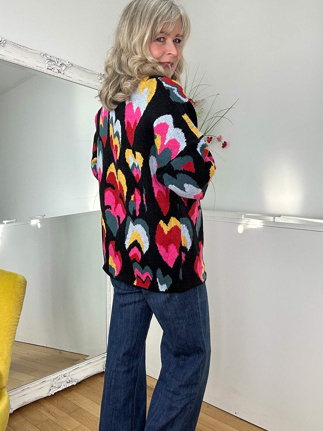 "A woman is standing with their back to the camera, wearing a colorful sweater with a heart pattern and blue jeans.