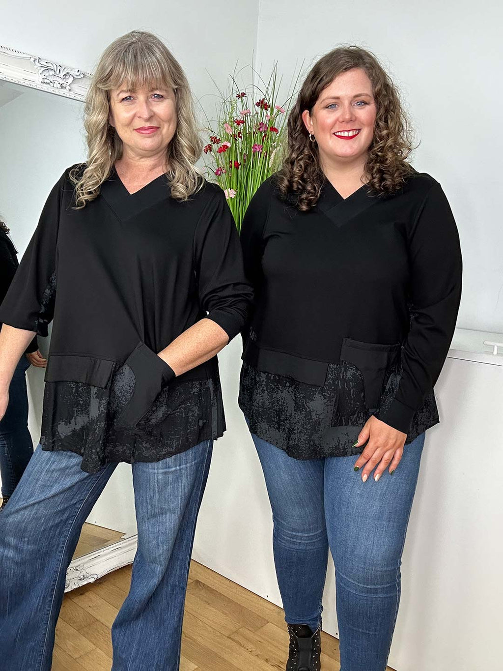 Two women are smiling indoors, showcasing their Helvig Tops with patterned accents. Made from a gorgeous, lasting fabric, the woman on the left flaunts her gray hair with jeans while the woman on the right shines in black pants or leggings.