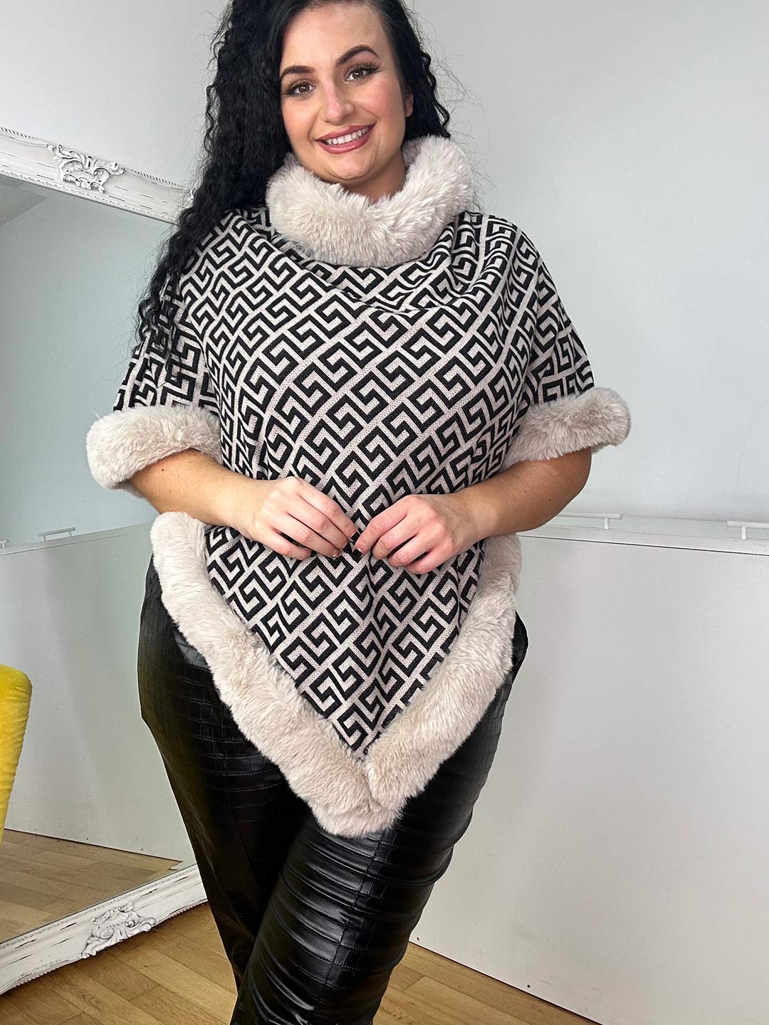 A person with long dark hair smiles while wearing the elegant Jill Poncho, featuring a versatile geometric pattern and fur trim, paired with black leather pants. Made from high-quality fabric, it drapes gracefully as they stand indoors against a light-colored wall and large mirror.