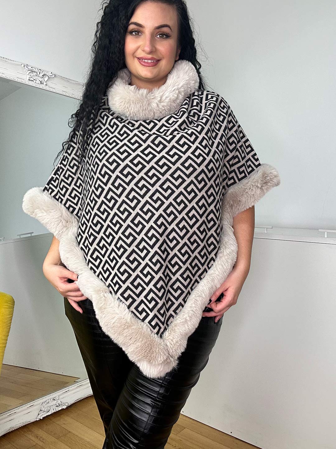 A person with long, curly hair is smiling while wearing the high-quality Jill Poncho with fur trim and black pants. The room features a light-colored wall, a mirror, and a yellow chair.