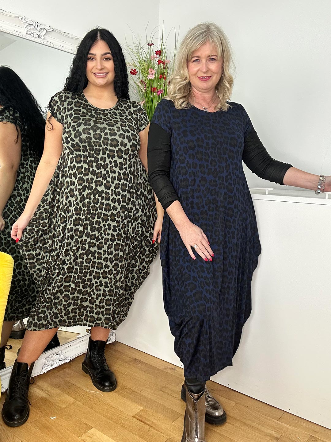  Two women wear leopard-print dresses, standing side by side, smiling in front of a mirror.