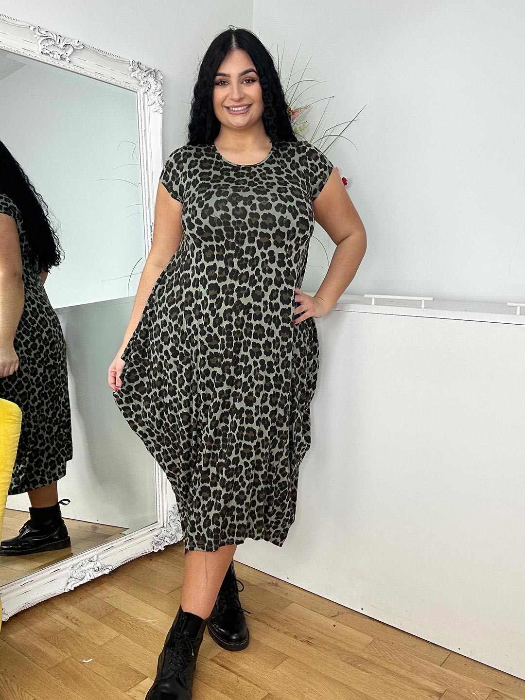 Smiling woman wears a leopard print dress, posing next to a mirror in a bright room.