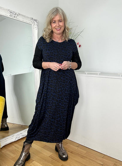 Woman in a navy leopard print dress and silver boots smiles in front of a mirror, with plants and decor in background.