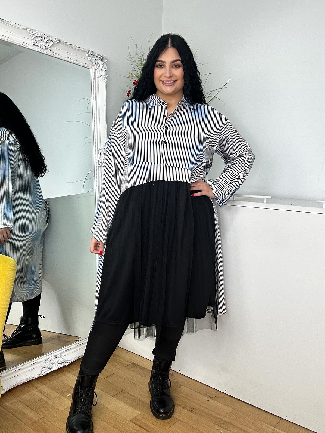 A woman wearing a grey shirt dress and black boots stands confidently, showcasing a stylish and modern outfit.