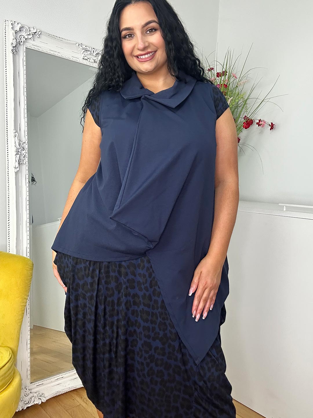 A smiling woman poses in a stylish dark outfit in front of a mirror, with plants and a yellow chair nearby
