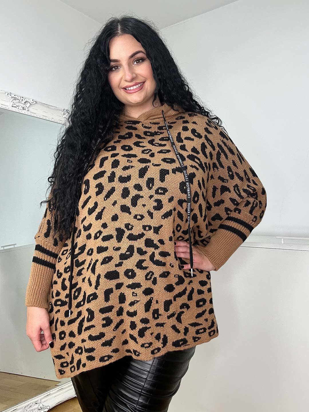 A person with long curly hair smiles warmly in a cozy Wendy Hoodie, featuring a one-size leopard print and hood, perfect for winter. Stylishly paired with black leather pants, they stand before a light-colored wall and framed mirror.