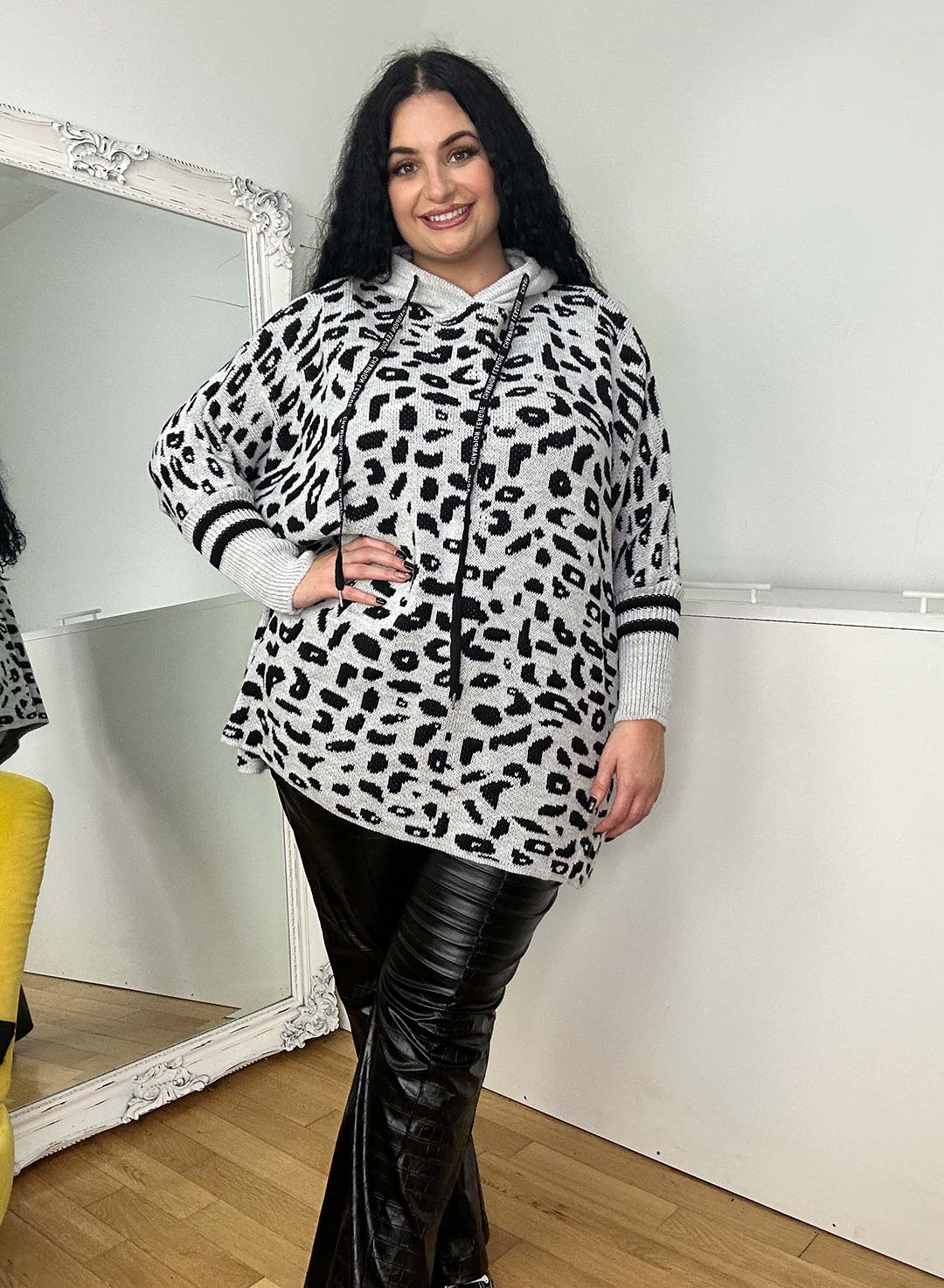 A person with long dark hair smiles, snug in their one-size Wendy Hoodie featuring a gray and black leopard print, paired with black leather pants. The mirror reflects their partial image, while a yellow chair adds warmth—perfect for cold winter days.