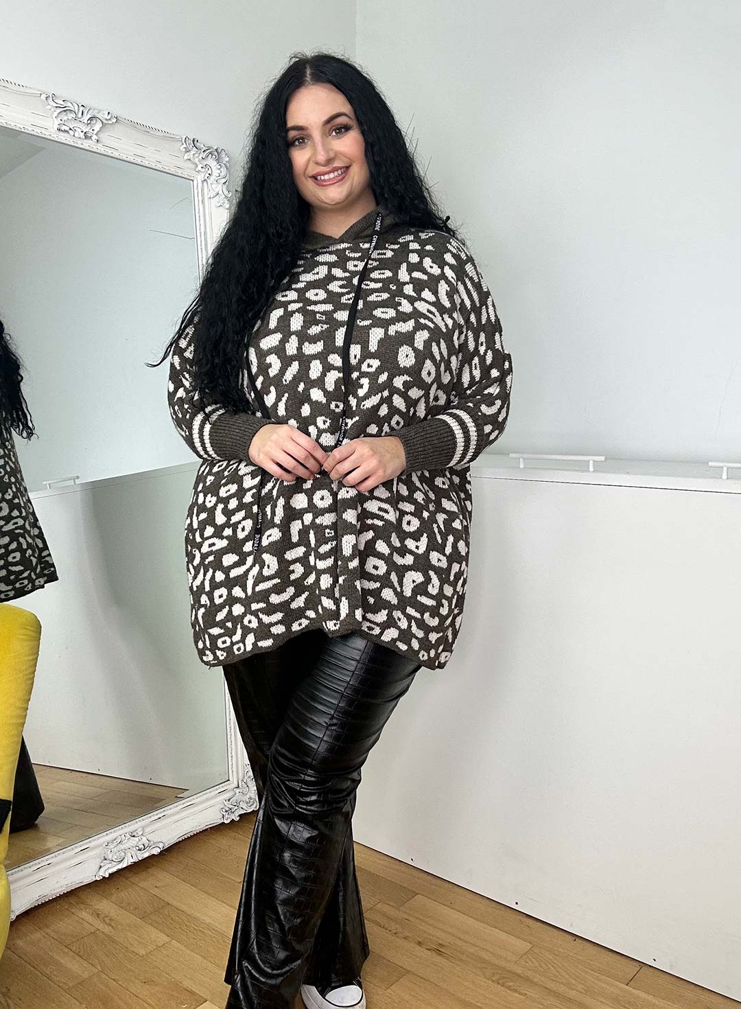 A woman with long black hair smiles while wearing a one-size patterned Wendy Hoodie and black pants. She stands before an ornate mirror on a light-colored wall, with a yellow chair partially visible to the left, perfectly capturing the cozy vibe of cold winter days.