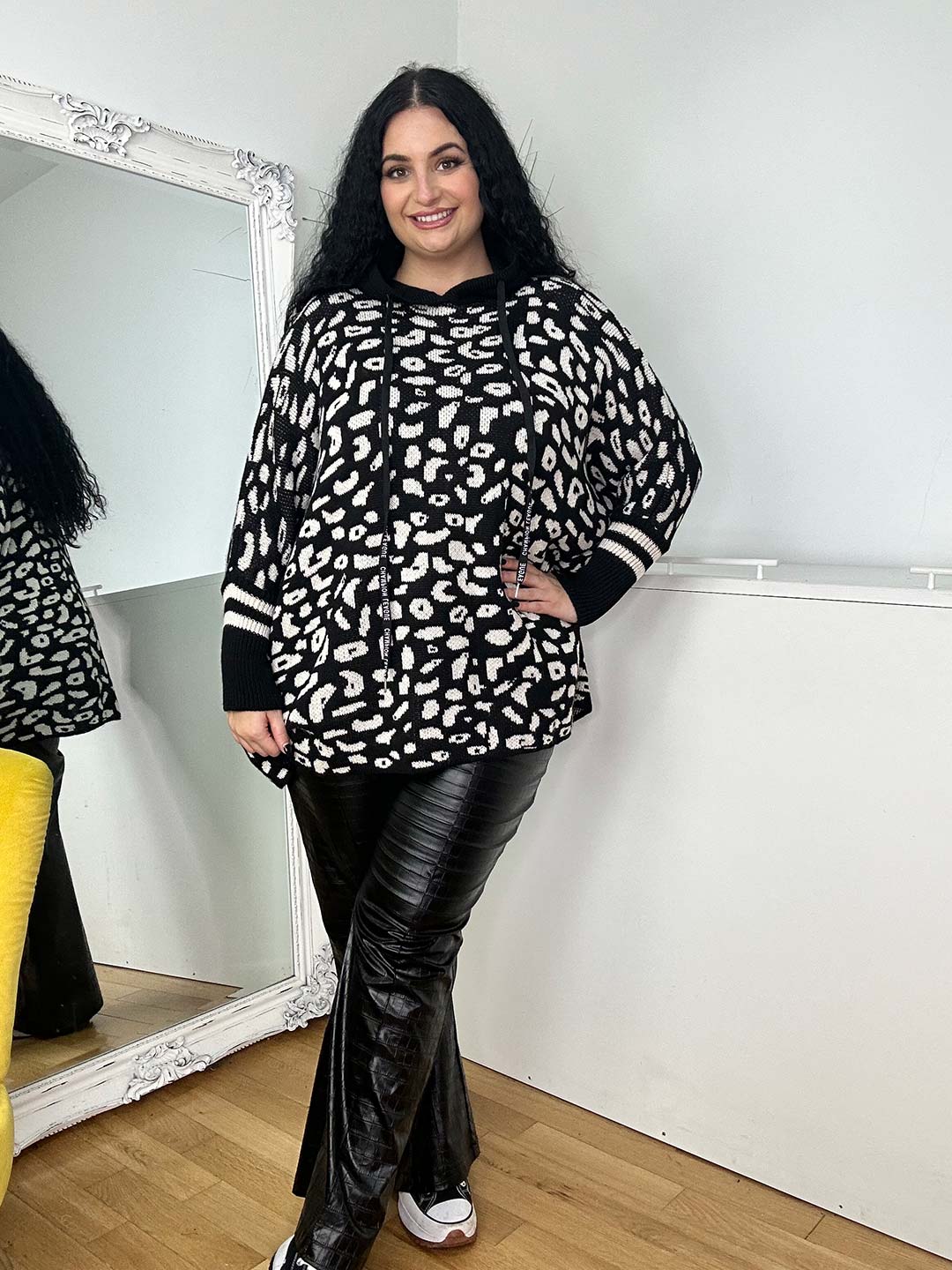 A person with long black hair stands indoors, wearing the Wendy Hoodie with a black and white pattern, ideal for cold days, paired with black leather pants. They smile and pose near a large white-framed mirror.