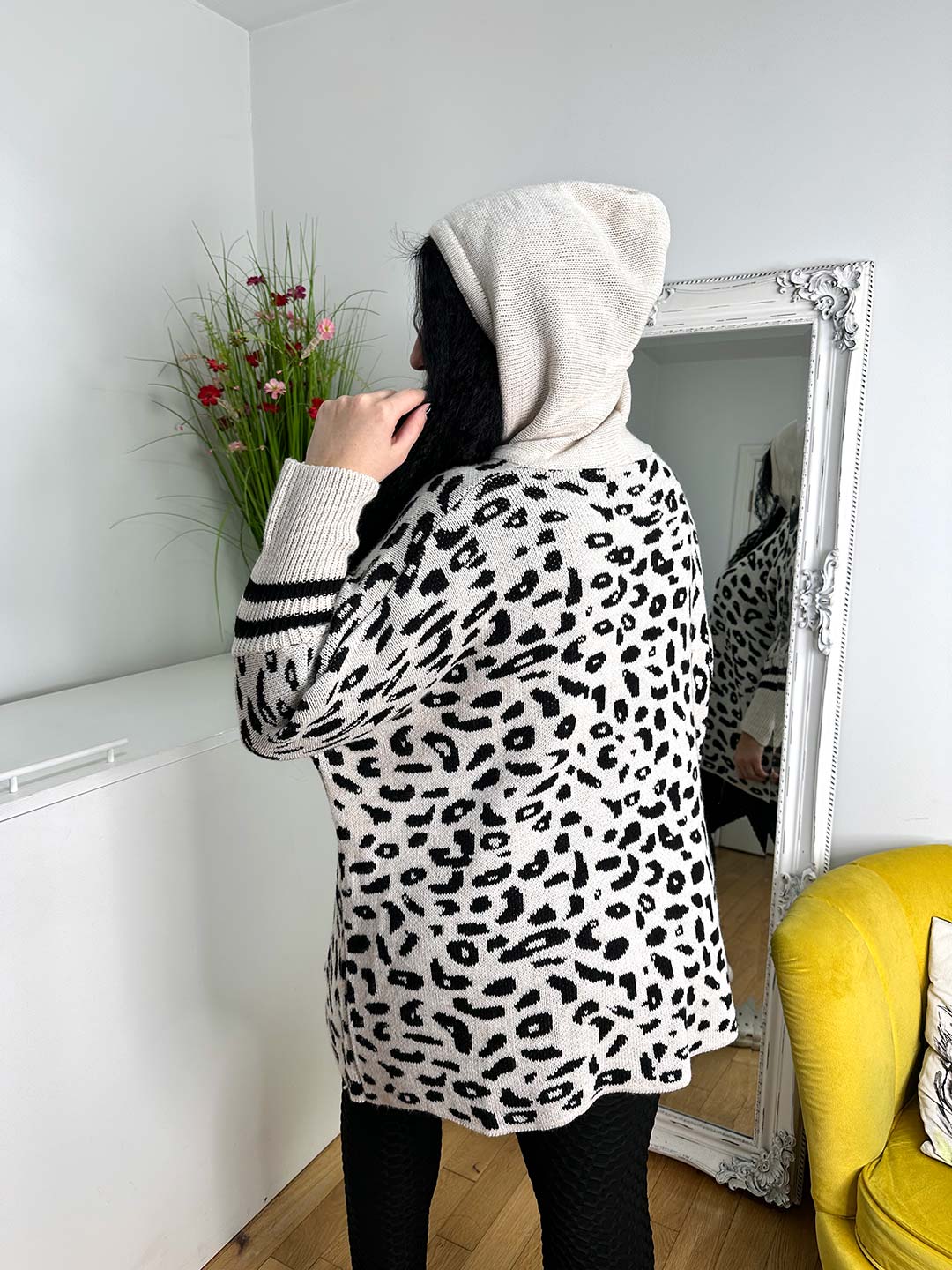 A person in a cozy, one-size leopard print sweater and the Wendy hoodie in beige stands facing a wall mirror. The room has a white cabinet, red-flowered potted plant, and yellow chair—ideal for chilly winter days.