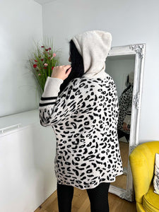 A person in a cozy, one-size leopard print sweater and the Wendy hoodie in beige stands facing a wall mirror. The room has a white cabinet, red-flowered potted plant, and yellow chair—ideal for chilly winter days.